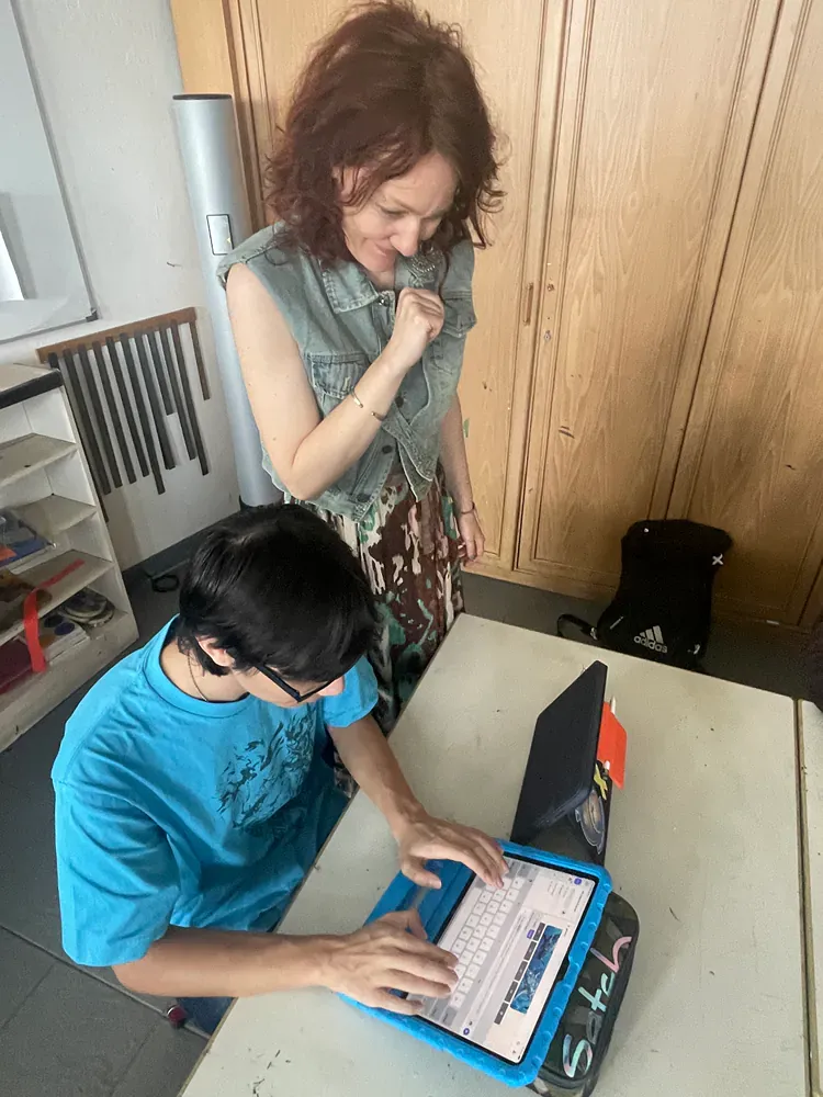 A woman is standing next to a boy using a laptop computer.
