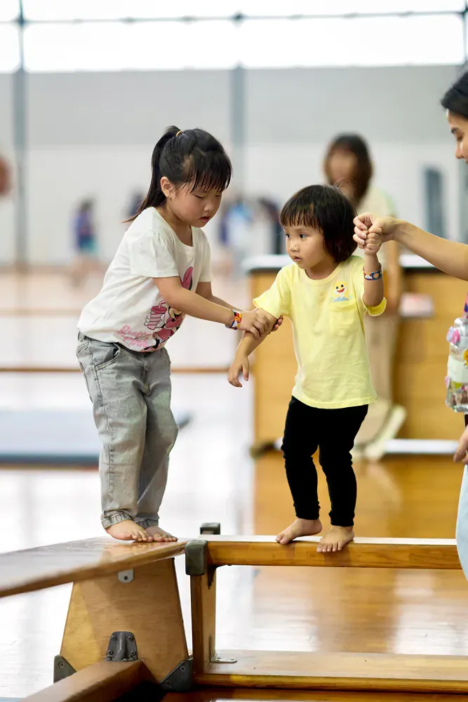 Two little girls are standing on a balance beam in a gym.