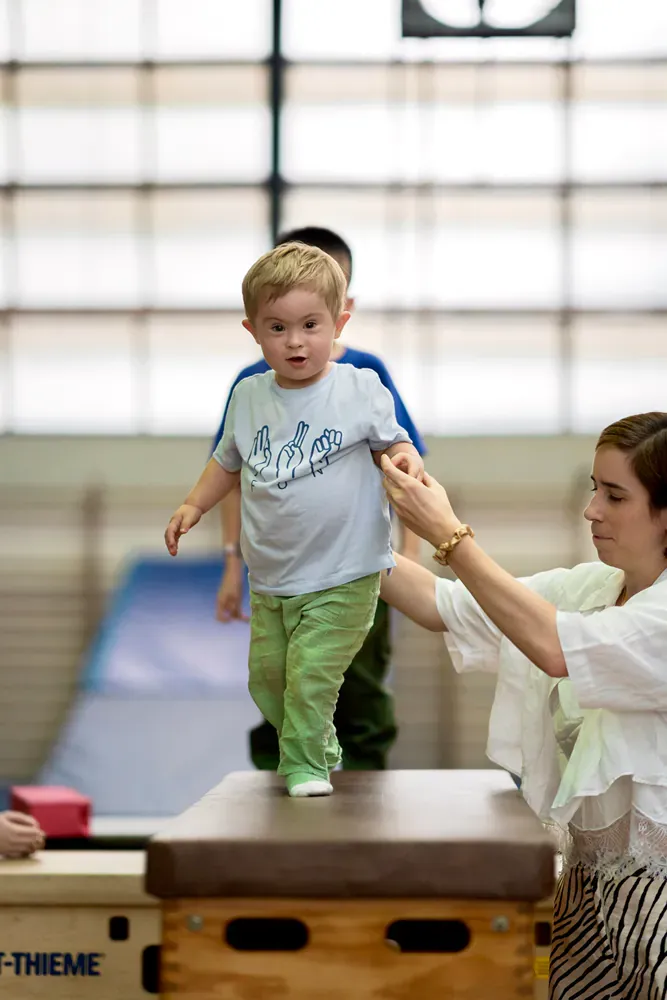 A woman is helping a young boy jump on a box in a gym.