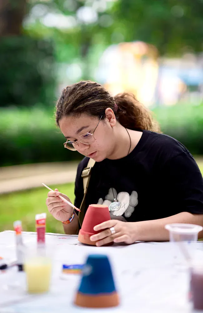 A woman is sitting at a table painting a pot with a brush.