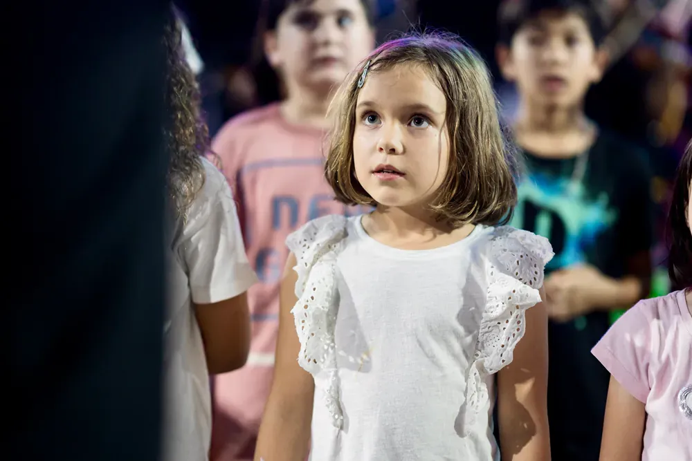 A little girl in a white shirt is standing in a crowd of children.