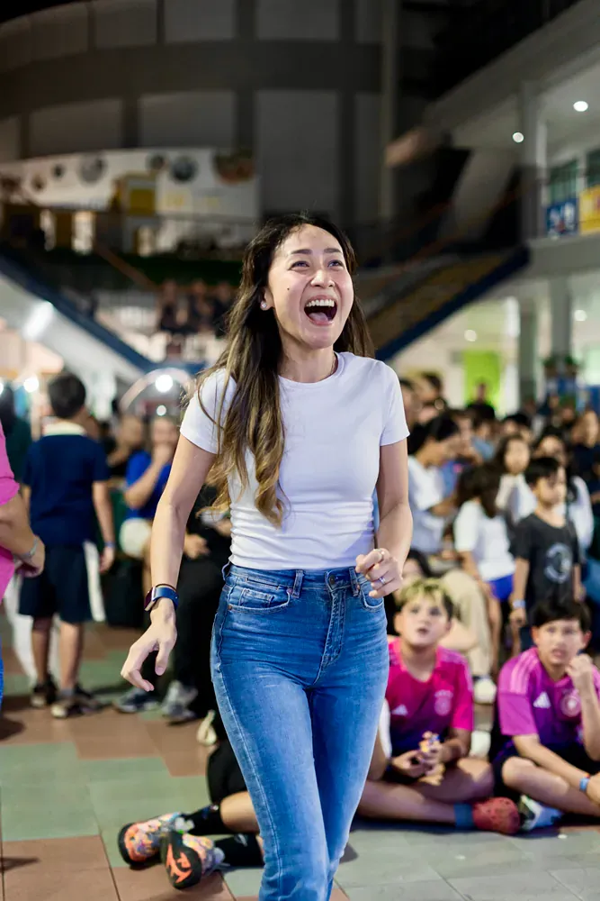 A woman in a white shirt and blue jeans is laughing in front of a crowd of people.
