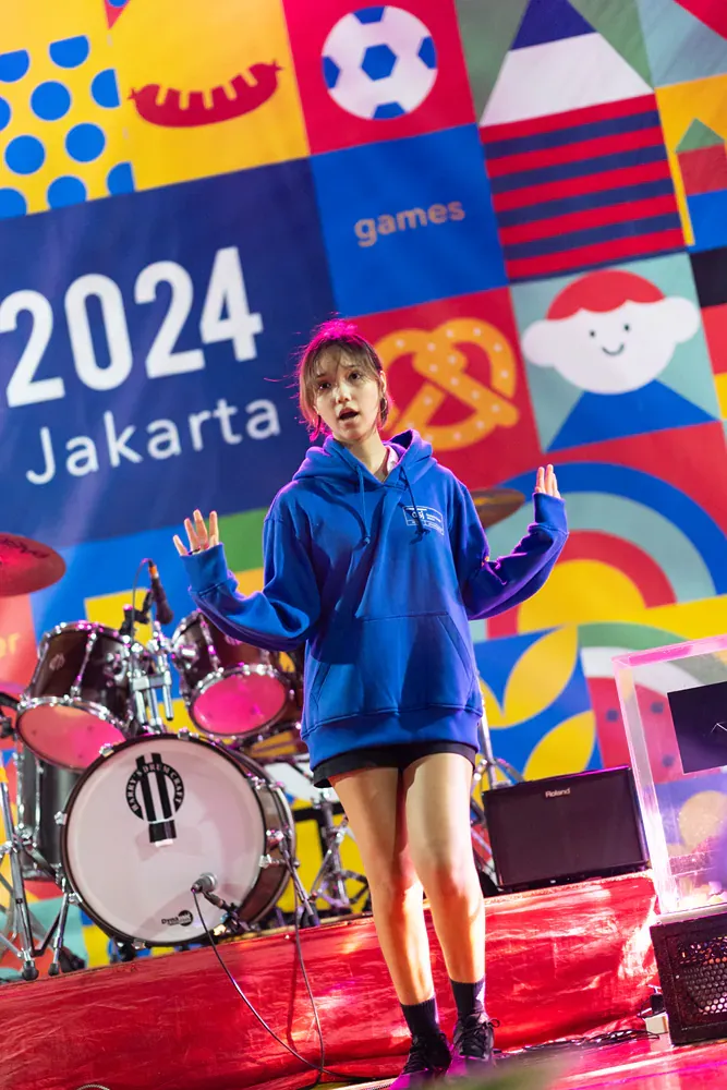 A woman in a blue hoodie is standing in front of a drum set.