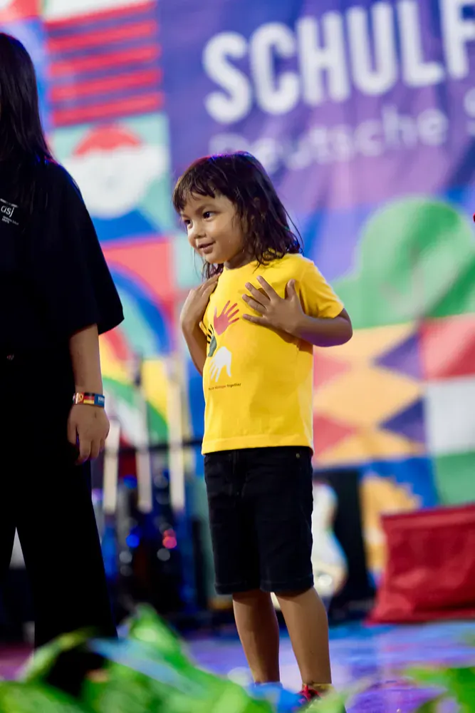 A little girl in a yellow shirt is standing in front of a schulf banner.