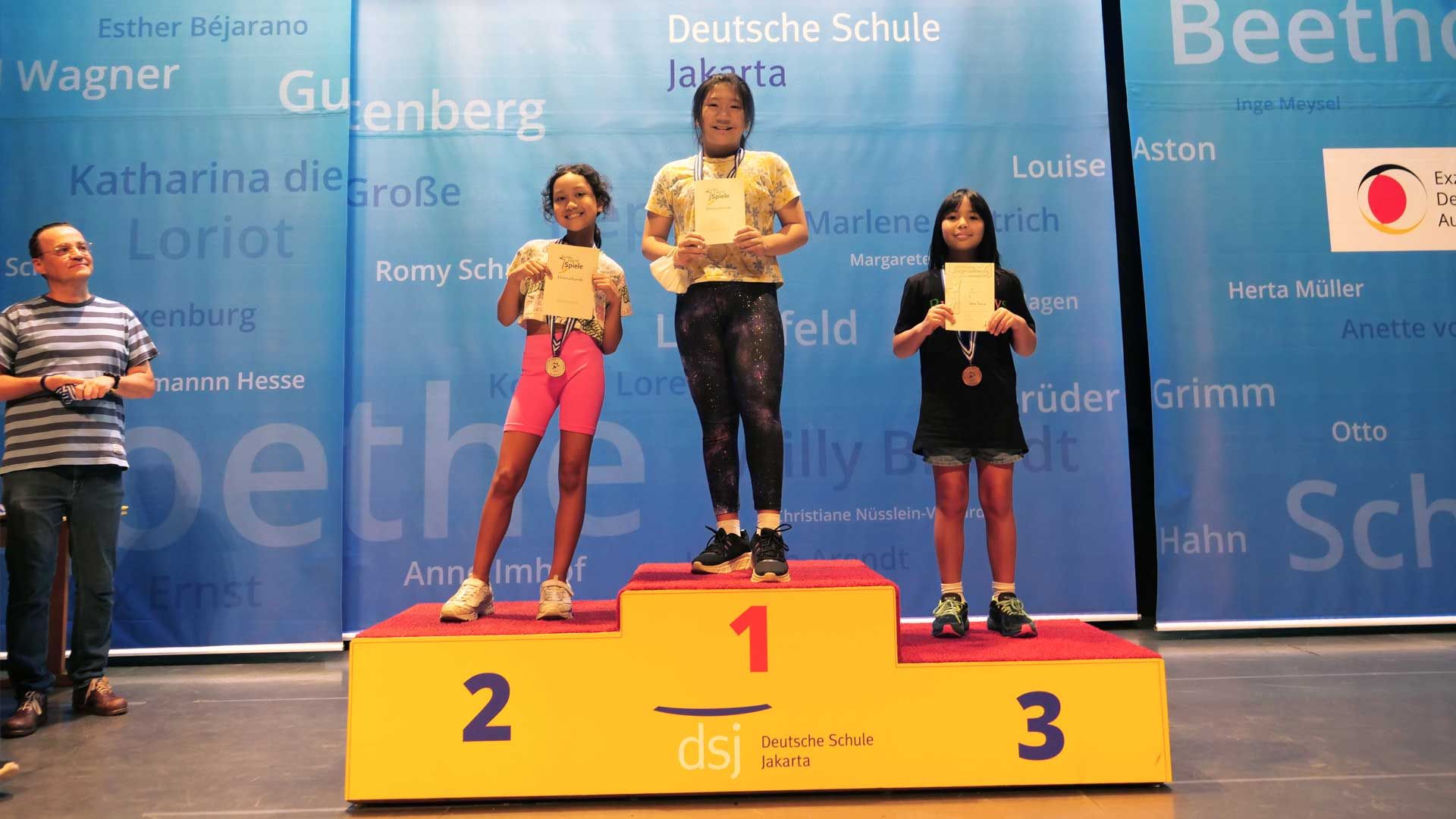 Three young girls are standing on a yellow podium with the number 1 on it