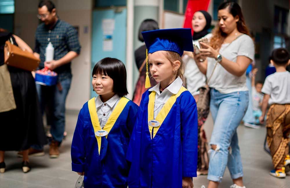 Two children in graduation caps and gowns are walking down a hallway.