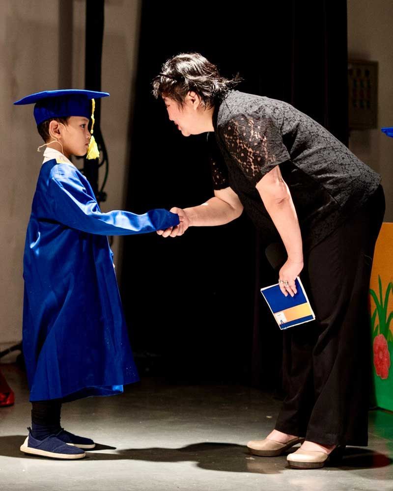 A little boy in a graduation cap and gown shakes hands with a woman