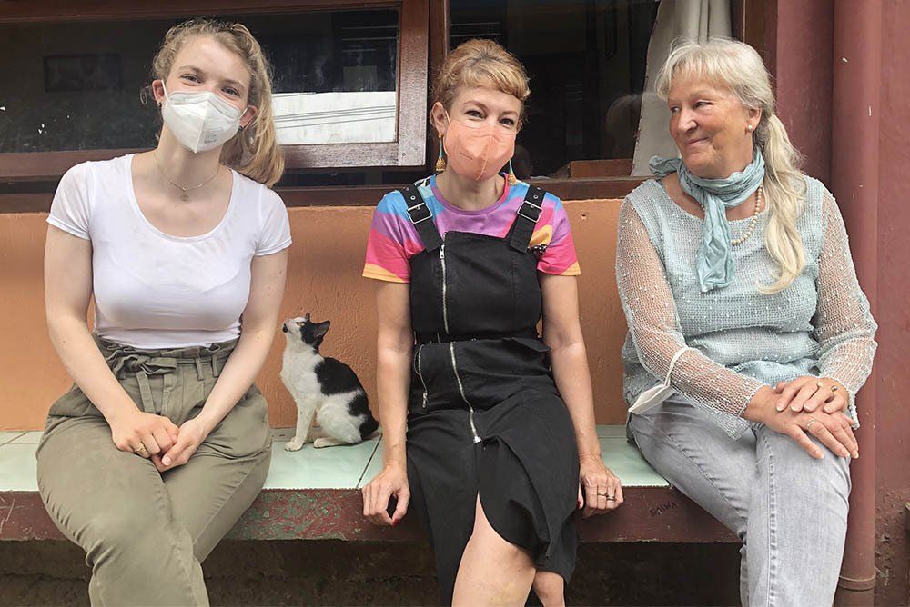 Three women wearing face masks are sitting on a bench with a cat.