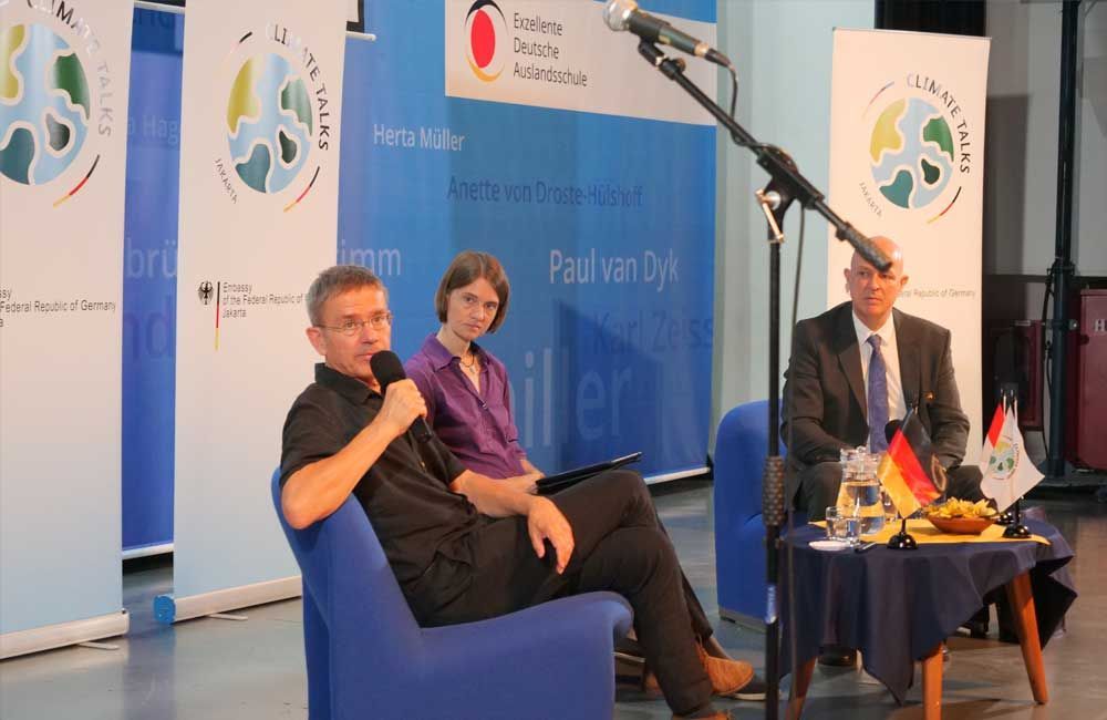 A man is sitting in a chair talking into a microphone while two other people sit at a table.