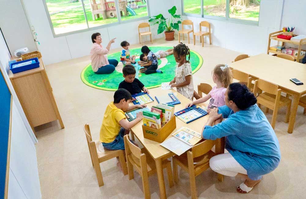 A group of children are sitting around a table in a classroom with a teacher.
