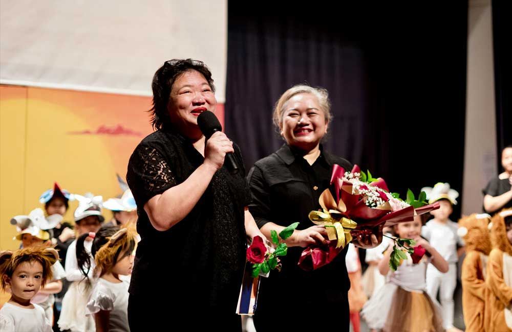 Two women are standing on a stage holding flowers and a microphone.