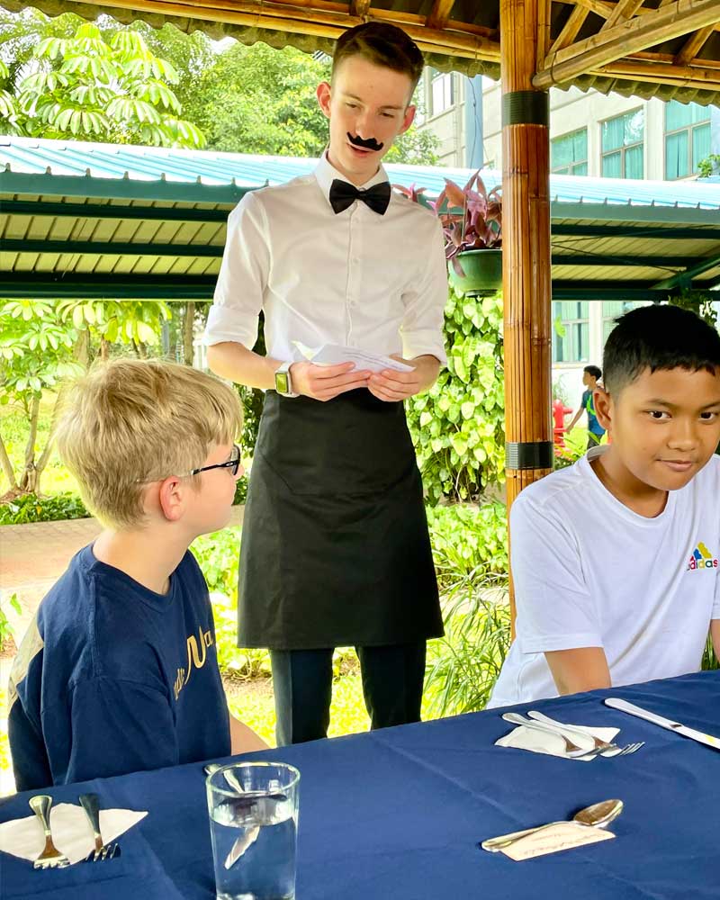 A man with a mustache is standing at a table with two boys.