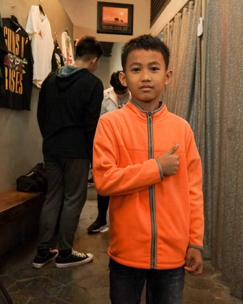 A young boy in an orange jacket is giving a thumbs up