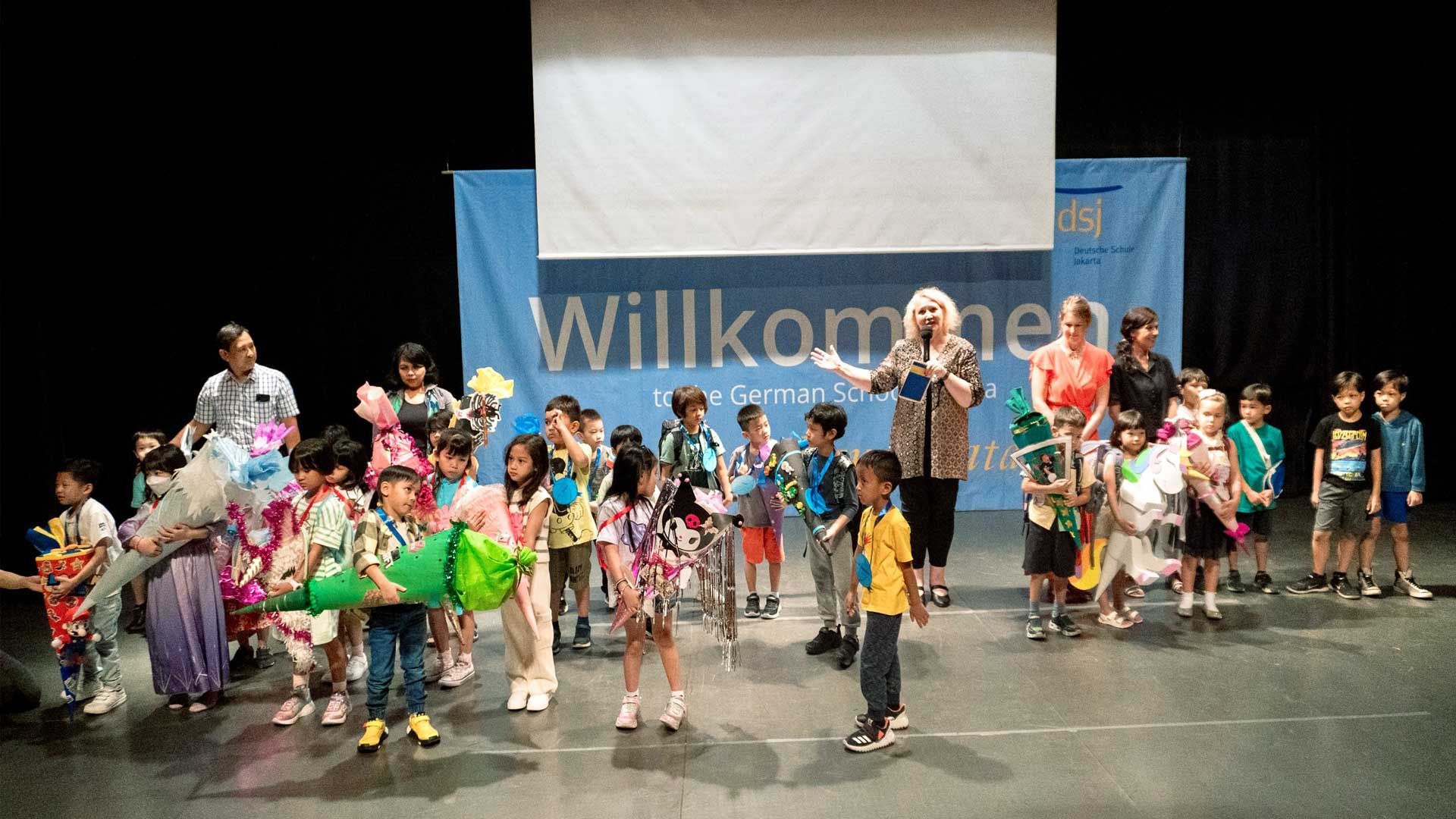 A group of children are standing on a stage in front of a sign that says willkommen