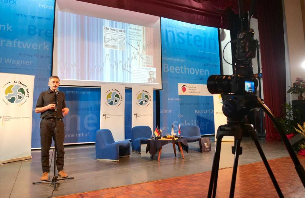A man is standing in front of a camera on a stage giving a presentation.