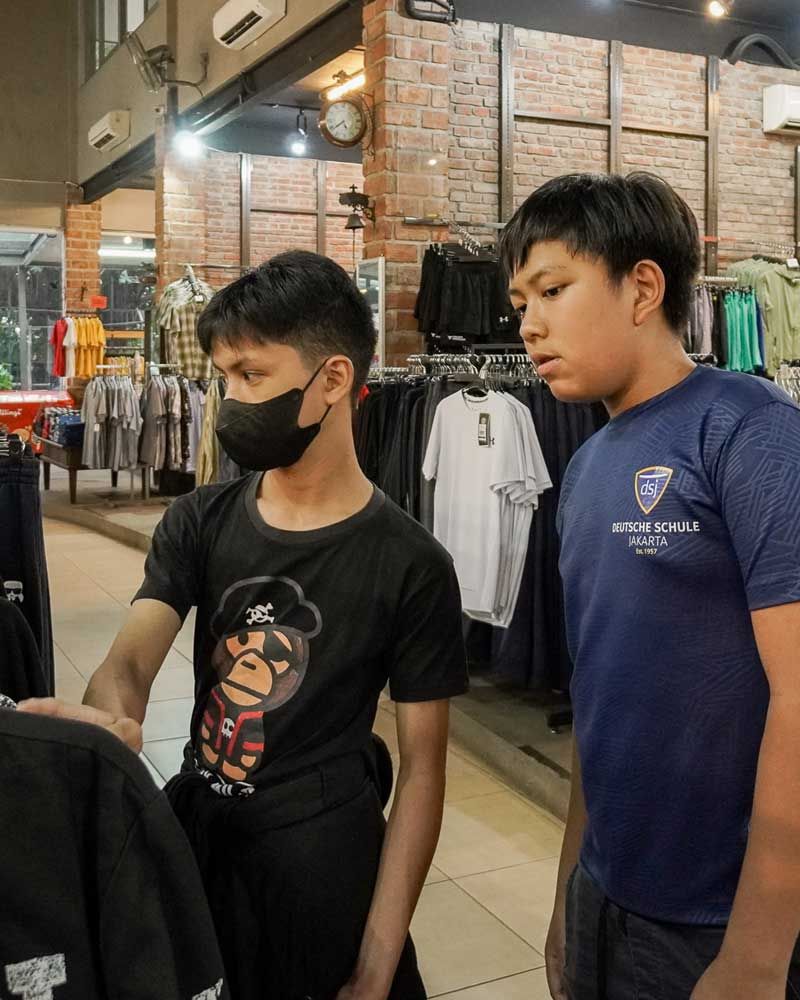 Two boys wearing masks are looking at clothes in a store.