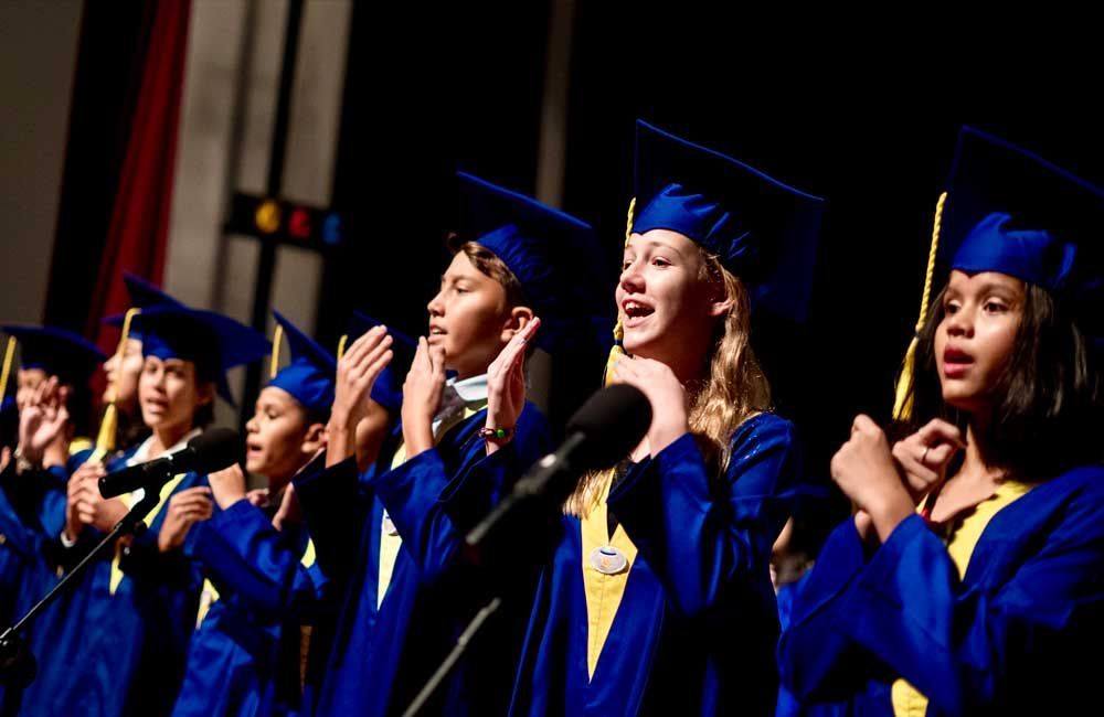 A group of graduates are singing into microphones on a stage.