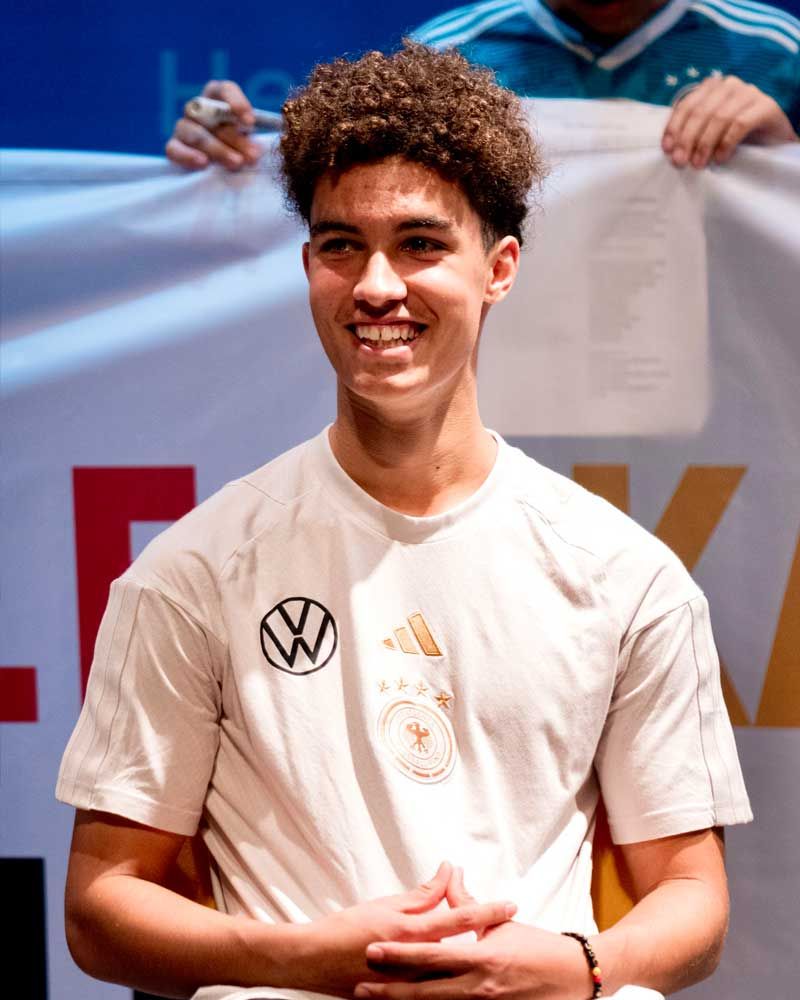 A young man with curly hair is wearing a white shirt with a vw logo on it.