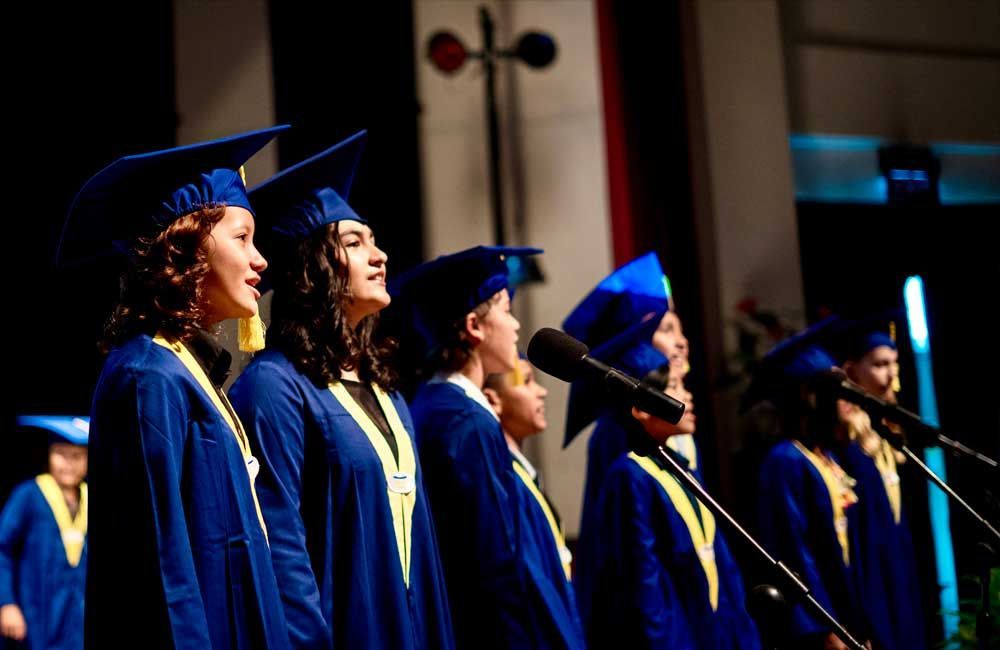 A group of graduates are singing into microphones on a stage.