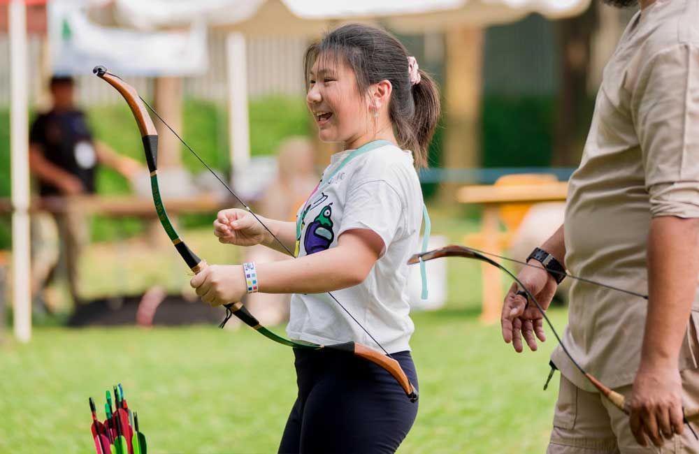 Young girl practicing archery outdoors with a bow and arrow, while an instructor stands beside her.