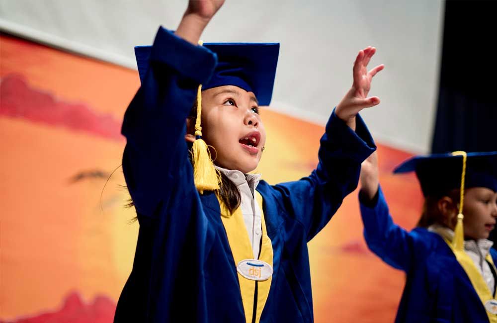 A young girl in a graduation cap and gown is raising her hands in the air.