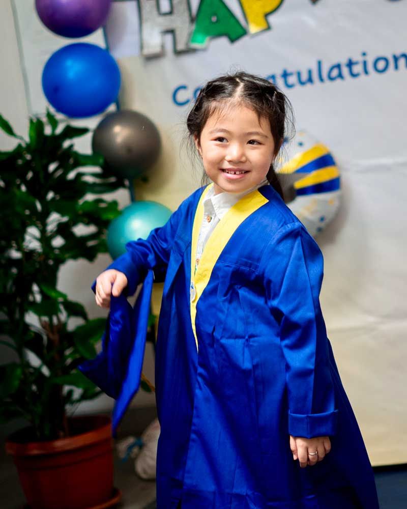A little girl in a blue graduation gown is standing in front of a sign that says congratulations.