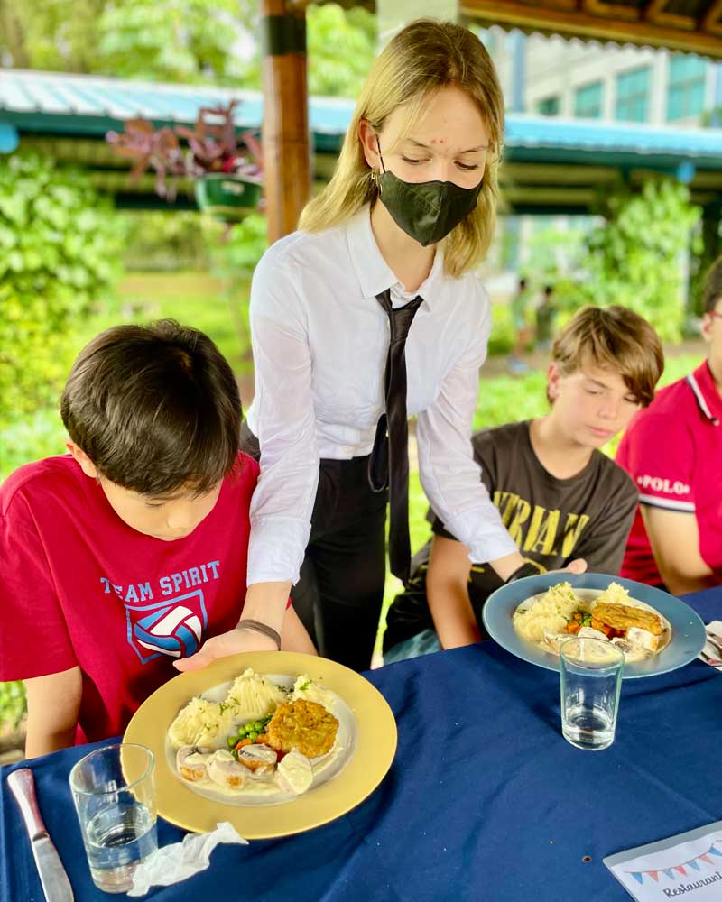 A woman wearing a mask is serving food to two children at a table.