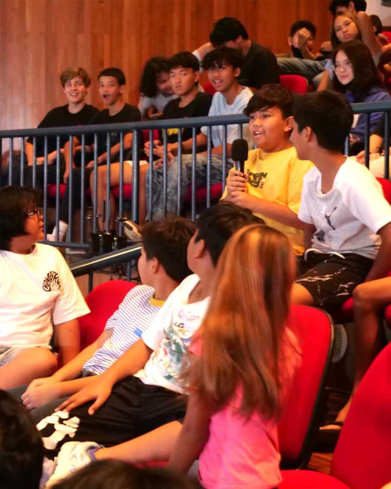 A group of people sitting in red chairs with a boy in a yellow shirt holding a microphone