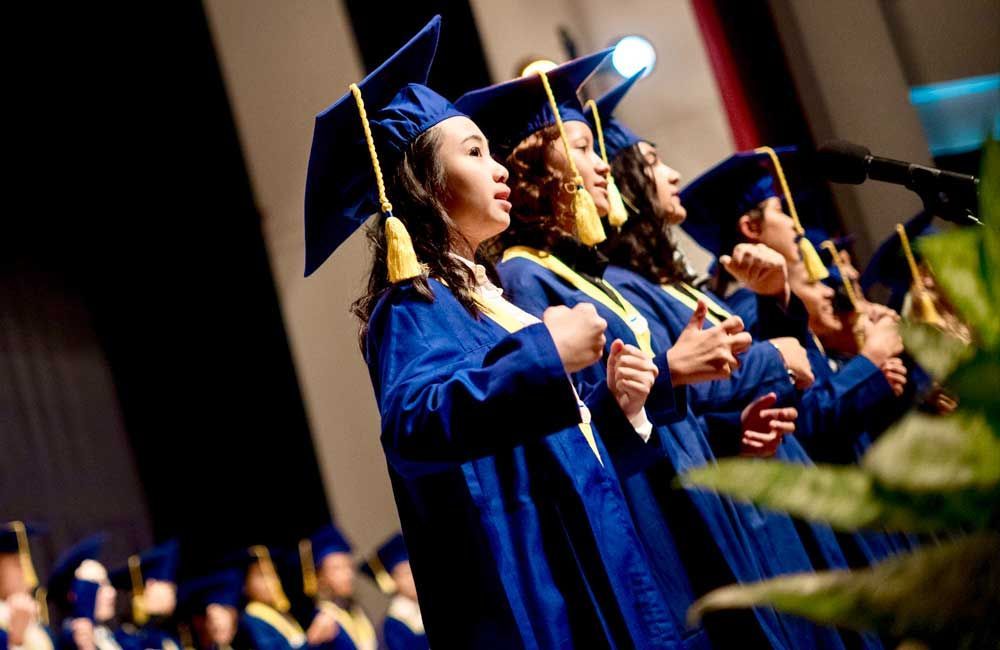 A group of graduates are singing into microphones on a stage.