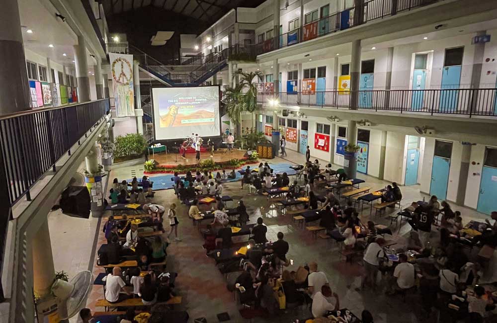 A large group of people are sitting at tables in front of a large screen.