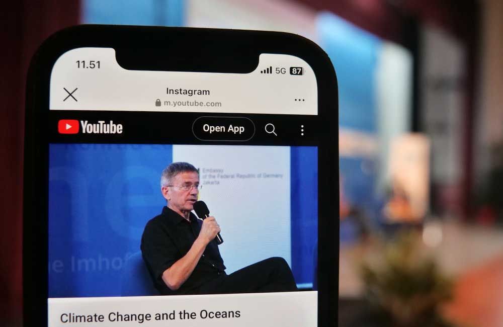 A cell phone is open to a youtube page with a man speaking into a microphone.
