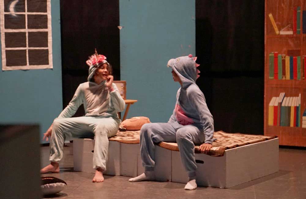 Two people in pajamas are sitting on a bed on a stage.