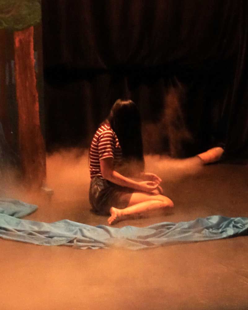 A woman is sitting on the floor on a stage with her legs crossed.