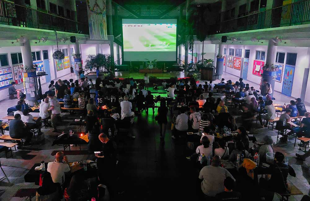 A large group of people are sitting at tables in front of a large screen.