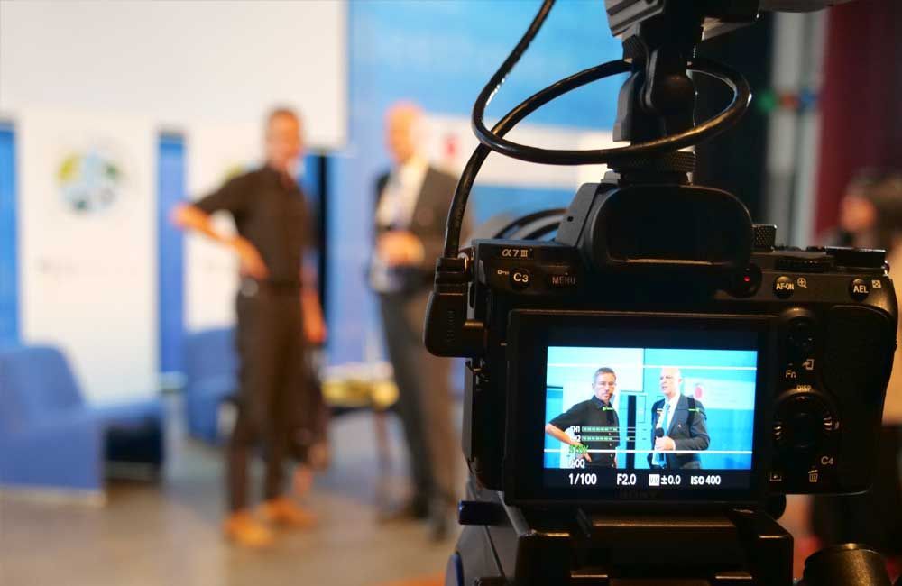 A camera is recording a man giving a presentation in front of a group of people.