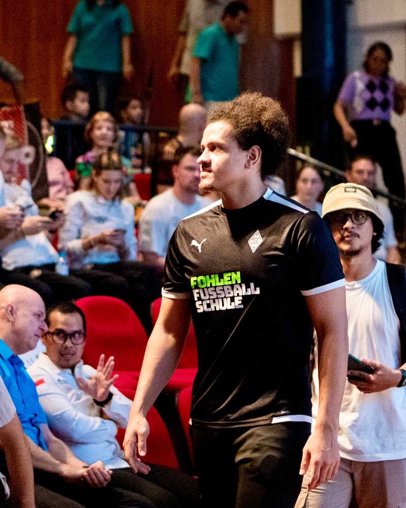 A man wearing a shirt that says fohlen fussball schule is walking through a crowd of people.