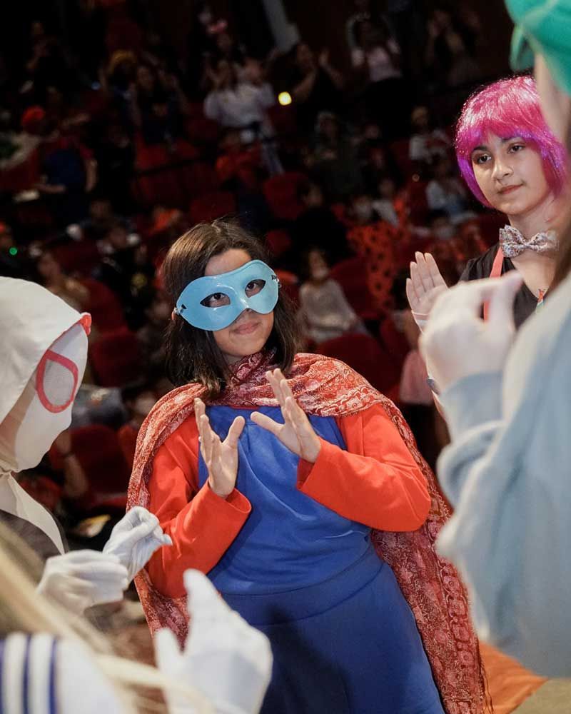 A woman wearing a mask and a cape is standing in front of a crowd.