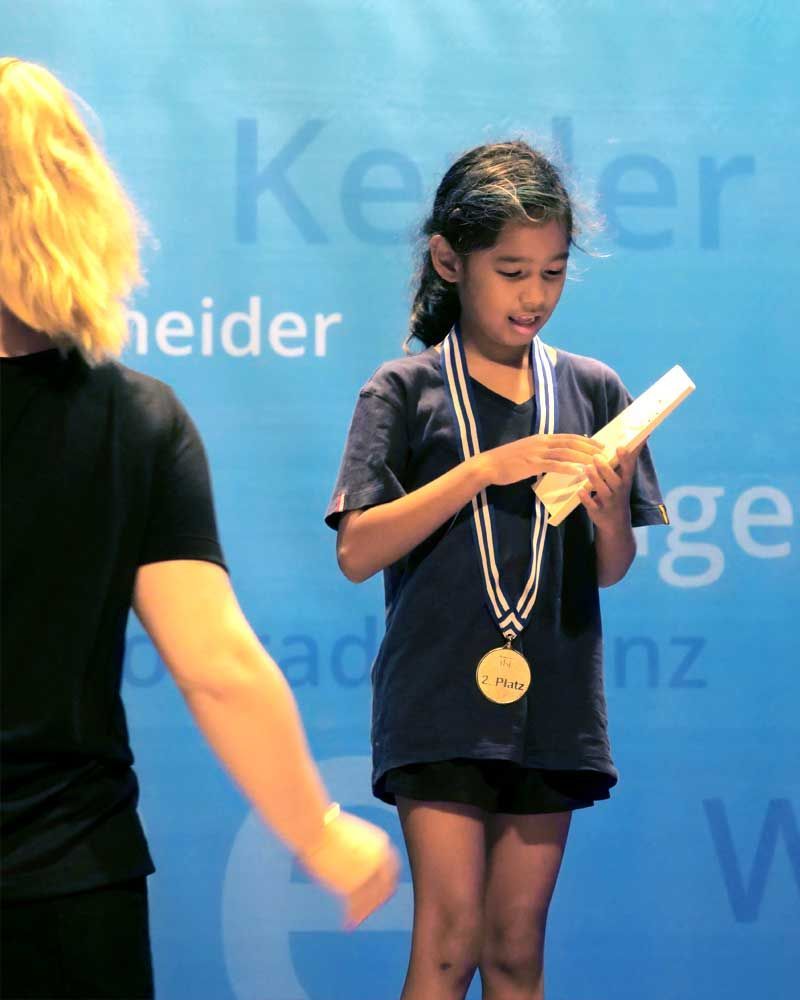 A little girl with a medal around her neck stands in front of a blue background that says heider