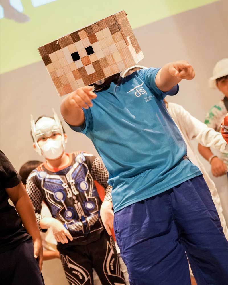 A boy in a blue shirt is wearing a minecraft mask on his head