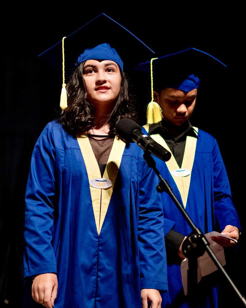 A woman in a graduation cap and gown stands in front of a microphone