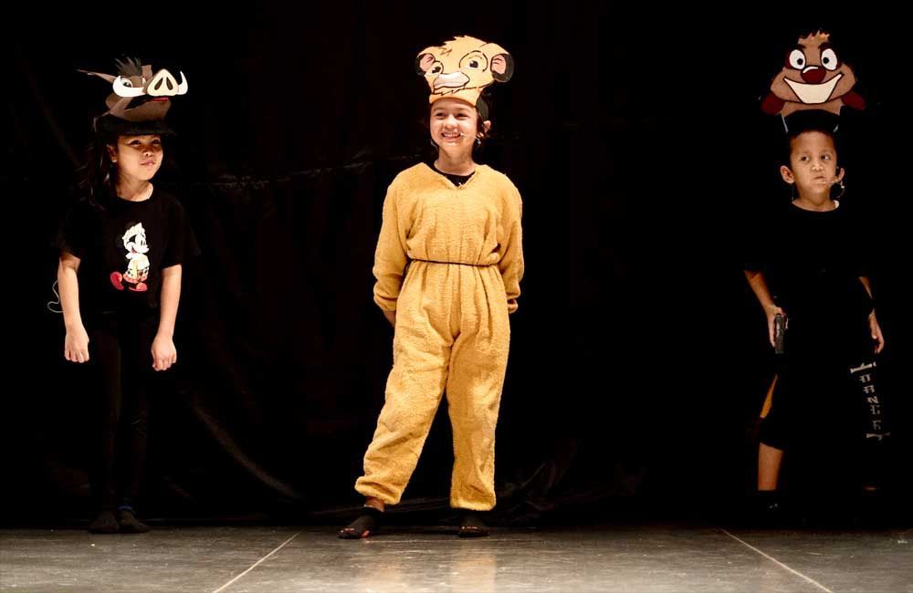 Three children are standing on a stage wearing lion king costumes.