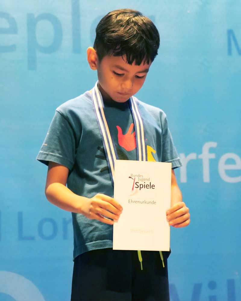 A young boy wearing a medal around his neck is holding a certificate