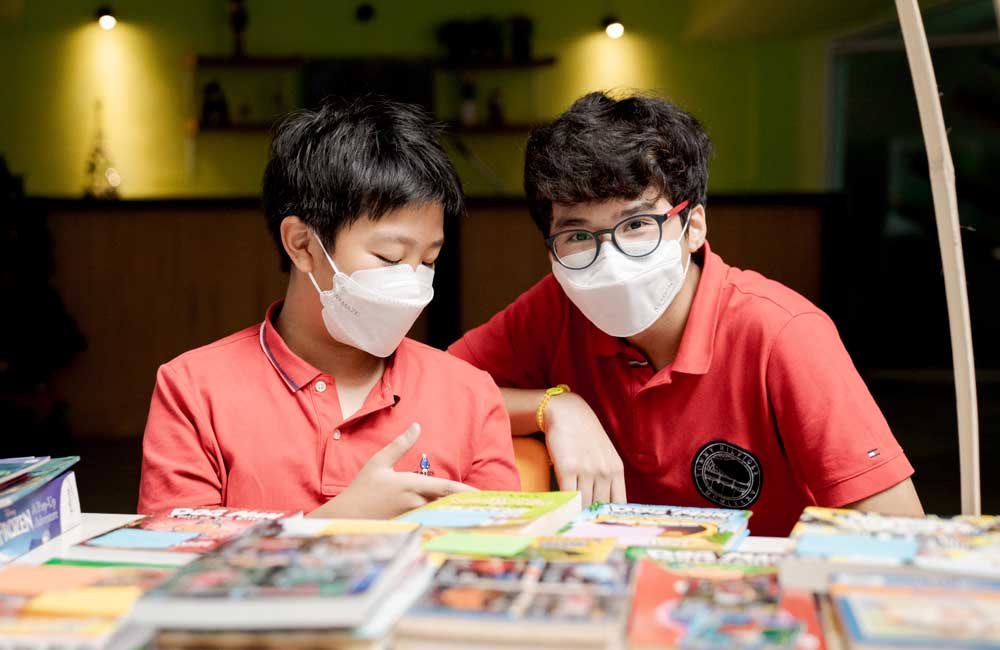 Two young boys wearing face masks are sitting at a table with books.