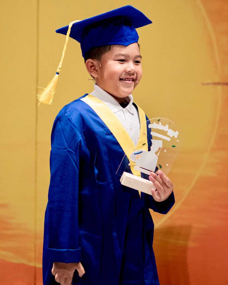 A young boy in a graduation cap and gown is holding a trophy.