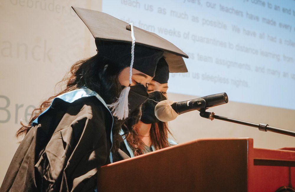 A woman in a graduation cap and gown is speaking into a microphone.
