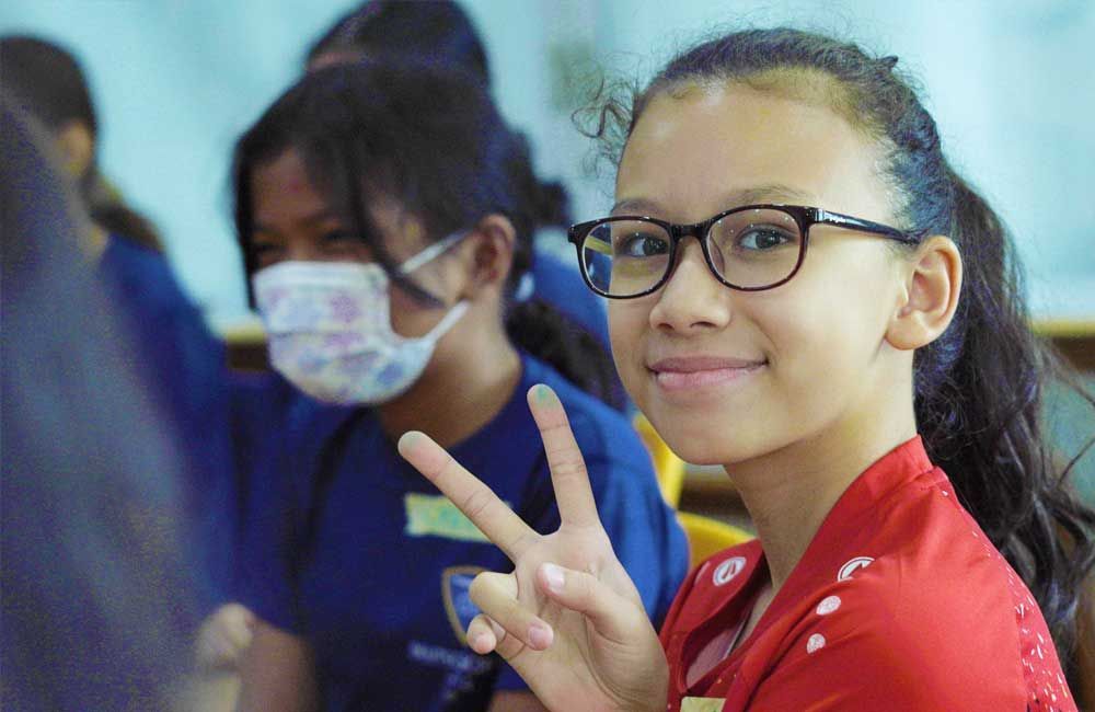 A girl wearing glasses and a red shirt is giving a peace sign in a classroom.
