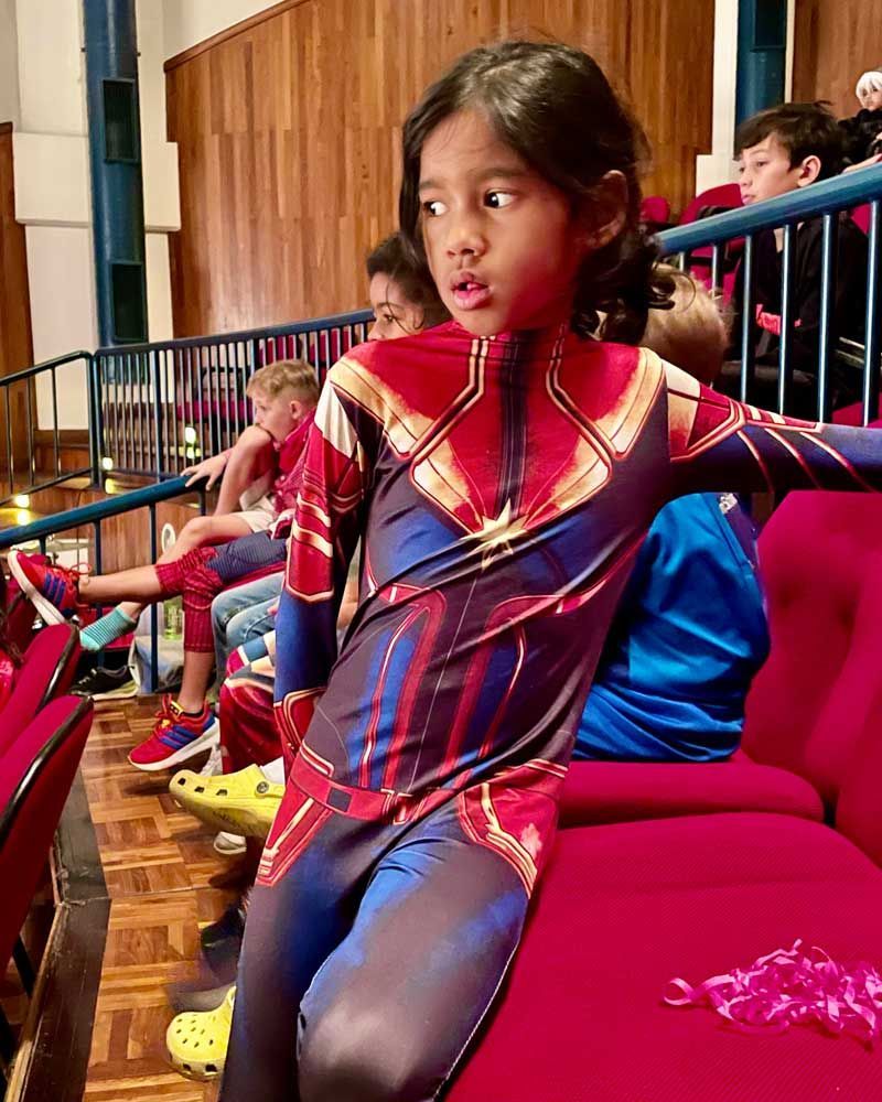 A young girl in a superhero costume is sitting in a row of red chairs.
