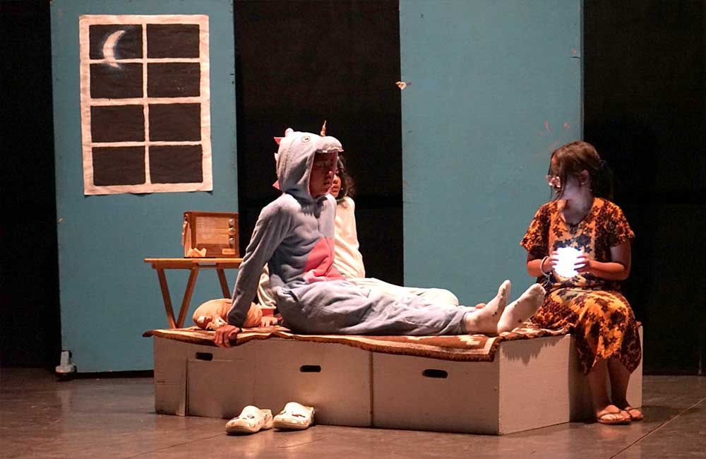 Two women are sitting on boxes on a stage in front of a window with a crescent moon on it