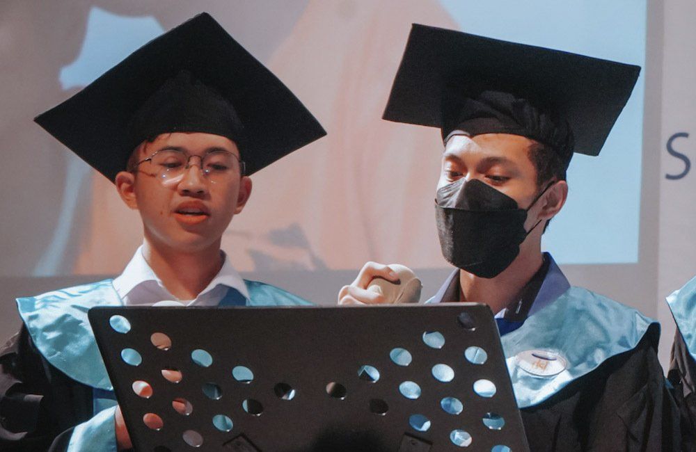 Two men in graduation caps and gowns are singing into a microphone.