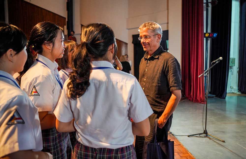 A man is standing in front of a microphone talking to a group of students.